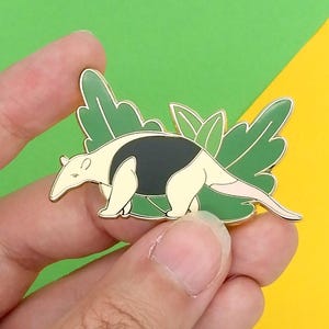 May include: A gold-coloured enamel pin featuring a cartoon anteater with black and white markings, standing on green leaves.