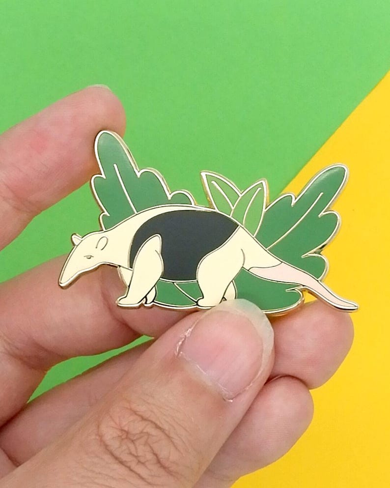 May include: A gold-coloured enamel pin featuring a cartoon anteater with black and white markings, standing on green leaves.