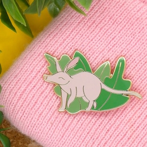May include: A gold-coloured enamel pin featuring a pink echidna with a long snout, standing on green leaves.