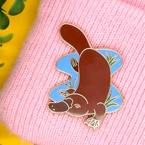 May include: A gold-tone enamel pin depicting a cartoon platypus swimming in blue water with green reeds. The platypus is brown with a white belly and has a small, pointed beak.