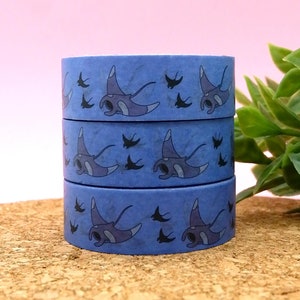 May include: Three rolls of blue washi tape featuring a pattern of manta rays and small birds. The tape is displayed on a cork surface.