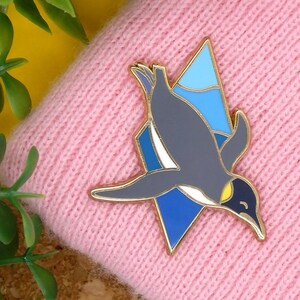 May include: A gold-tone enamel pin featuring a penguin diving into blue and white water. The penguin is grey with a white belly and a yellow beak.