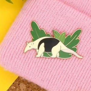 May include: A gold-coloured enamel pin featuring an anteater with black and white markings, walking through green leaves. The pin is on a pink knitted beanie.