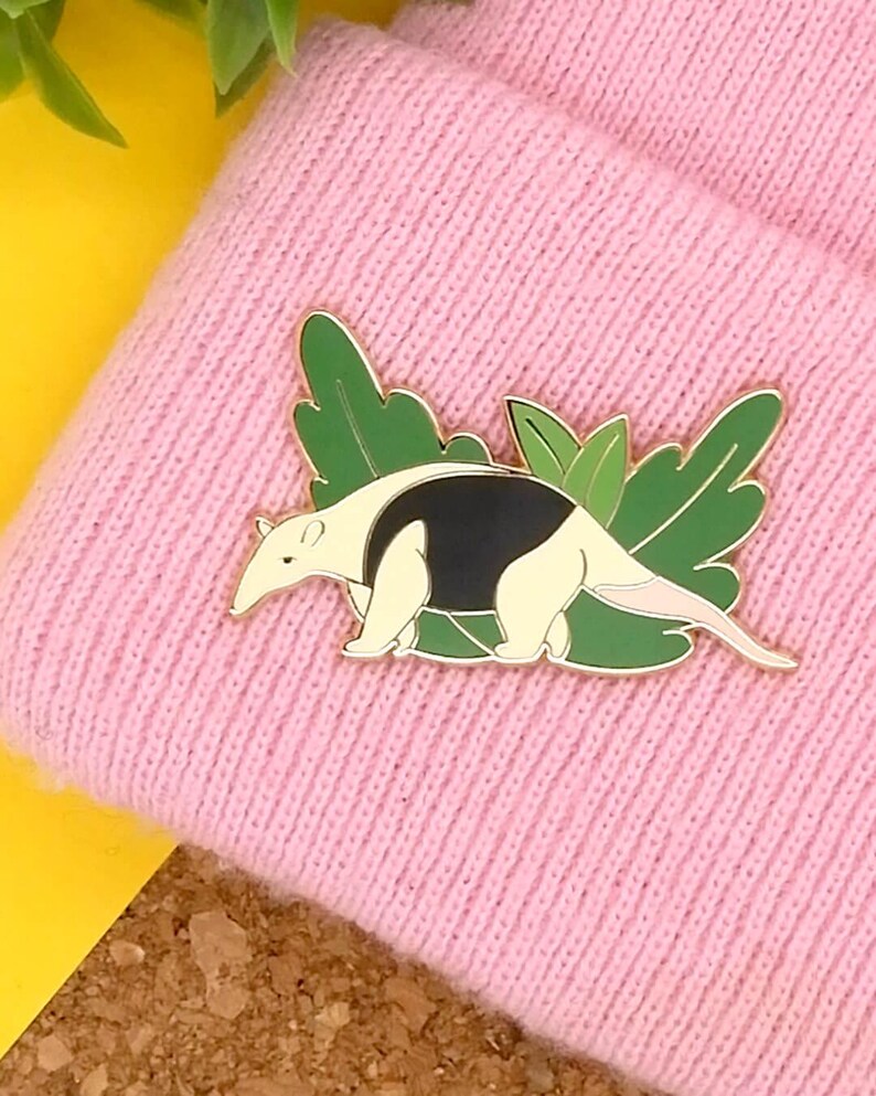 May include: A gold-coloured enamel pin featuring an anteater with black and white markings, walking through green leaves. The pin is on a pink knitted beanie.