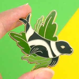 May include: A gold-tone enamel pin featuring a black and white stylized fish with green leafy wings. The fish has a white stripe down its body and a green leafy design around its head.