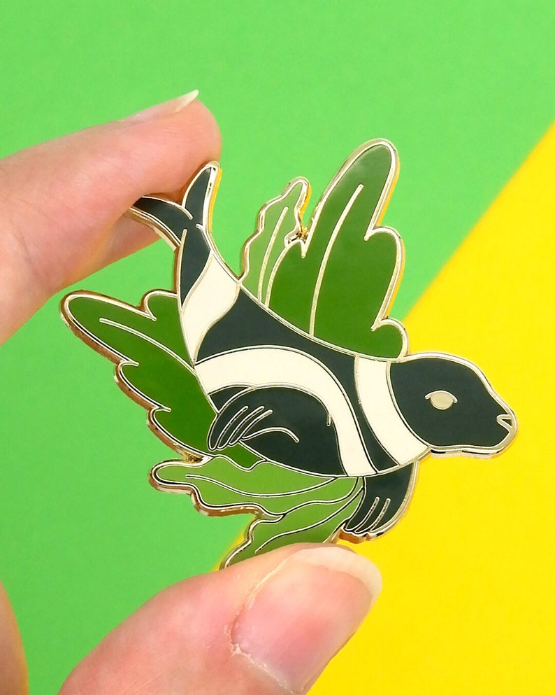 May include: A gold-tone enamel pin featuring a black and white stylized fish with green leafy wings. The fish has a white stripe down its body and a green leafy design around its head.