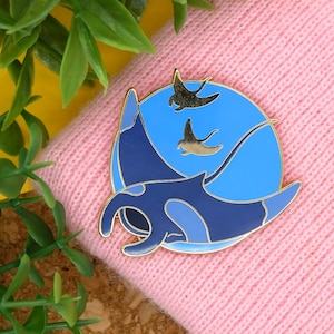 May include: A gold-tone enamel pin featuring a blue and black manta ray with three smaller black manta rays in the background. The pin is set against a pink knitted fabric.