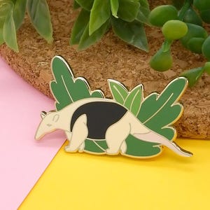 May include: A gold-coloured enamel pin featuring a cartoon anteater with black and white markings, surrounded by green leaves.