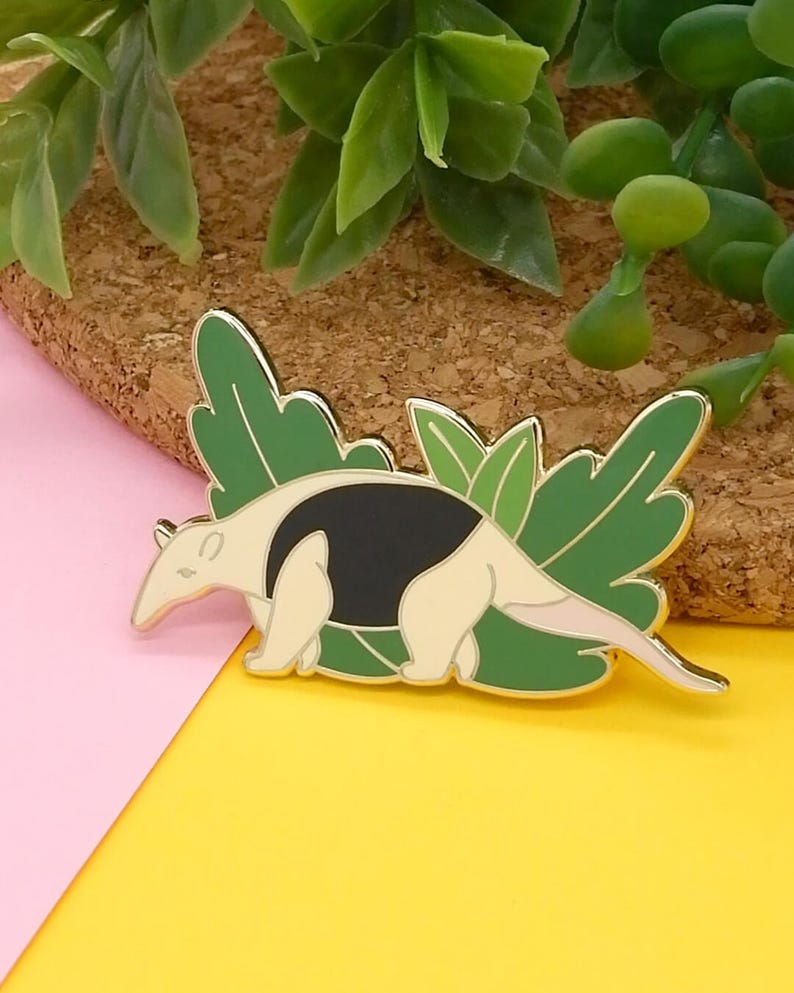 May include: A gold-coloured enamel pin featuring a cartoon anteater with black and white markings, surrounded by green leaves.