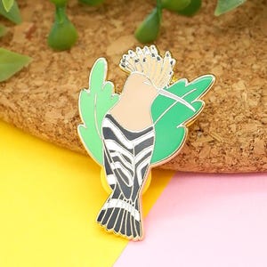 May include: Gold-tone enamel pin featuring a hoopoe bird with black and white stripes, a brown head, and a crest of feathers. The bird is perched on a green leaf.