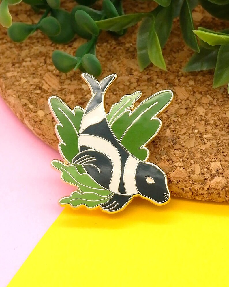 May include: Gold-tone enamel pin featuring a black and white striped fish swimming through green seaweed.