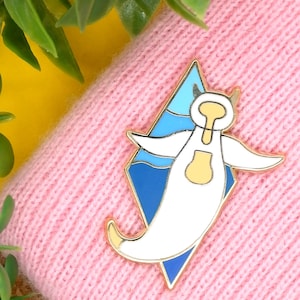 May include: A white enamel pin with a gold outline depicting a cartoon ghost wearing a yellow life preserver and floating in a blue and white ocean. The pin is shaped like a diamond and is attached to a pink knitted fabric.