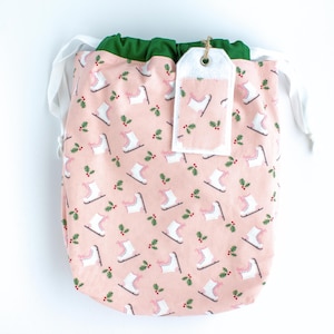 May include: A pink fabric gift bag with a white drawstring and a white tag. The bag is decorated with a repeating pattern of pink ice skates and green holly sprigs.