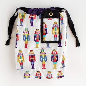 Puede incluir: Una bolsa blanca con cordón y un patrón repetido de figuras de cascanueces coloridas. La bolsa tiene un forro de lunares morados, cordones negros y una etiqueta negra con una arandela dorada.