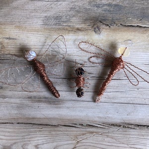May include: Three wire sculptures of dragonflies, one with a white bead for a head, one with a light yellow bead for a head, and one with a black bead for a head. The sculptures are on a wooden surface.