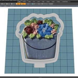 May include: A white cookie cutter in the shape of a bucket with a bouquet of flowers. The flowers are pink, blue, and brown with green leaves.