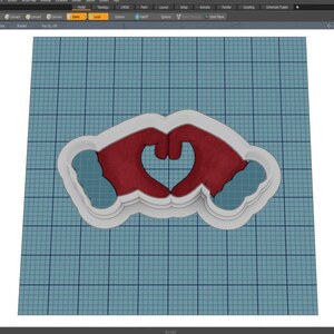 May include: A white plastic cookie cutter shaped like two hands forming a heart. The hands are red and the heart is outlined in black.