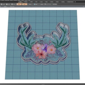 May include: A 3D model of a cookie cutter in the shape of a floral wreath with antlers. The wreath is decorated with pink, purple, and green flowers. The cookie cutter is made of silver metal.