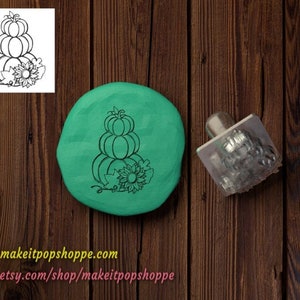 May include: A close-up of a green polymer clay imprint of a stack of three pumpkins with a sunflower and leaves. A metal stamp with the same design is also shown.