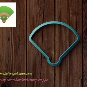 May include: A teal blue plastic cookie cutter shaped like a baseball field. The cookie cutter is on a brown wooden background.