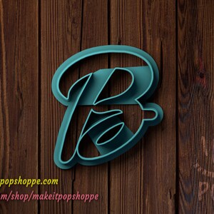 May include: A teal blue plastic cookie cutter in the shape of the letter "B" with a cursive font. The cutter is on a brown wooden background.