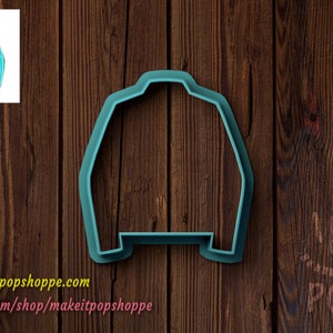 May include: A teal blue plastic cookie cutter shaped like a sweater with the word "Bride" in pink lettering on the front. The cookie cutter is on a brown wooden surface.