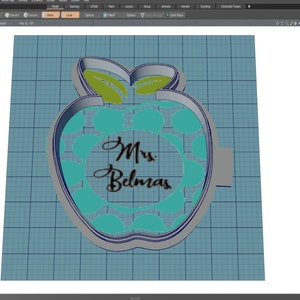 May include: A grey 3D model of a cookie cutter shaped like an apple with green leaves. The apple has a circle in the centre with the text "Mrs. Belmas" written in black.