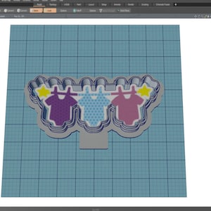 May include: A 3D model of a cookie cutter shaped like a clothesline with three baby onesies hanging on it. The onesies are purple, blue with white polka dots, and pink. Each onesie has a yellow star above it.