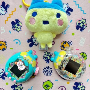 Puede incluir: Un llavero de peluche amarillo y azul con un anillo amarillo, y dos juguetes electrónicos portátiles. Uno es turquesa con un botón rosa y el otro es transparente con detalles amarillos. El fondo es rosa con personajes de dibujos animados.