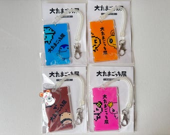 May include: Four colorful ID card holders with pixelated character designs and the text "大たまごっち展". Each holder has a clear plastic case, a coiled lanyard, and a metal clasp. Colors include blue, orange, burgundy, and pink.