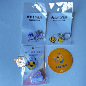 May include: Assortment of Tamagotchi merchandise, including keychains and hair clips. The keychains feature pixelated and cartoon character designs. The hair clips are in a clear plastic package with Japanese text. A yellow circular sticker with a character illustration is also visible.