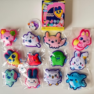 May include: A collection of colorful Tamagotchi character patches in individual plastic packaging. The patches feature various cute animal designs in pink, blue, yellow, and green. A box of Tamagotchi products and a round sticker are also visible.