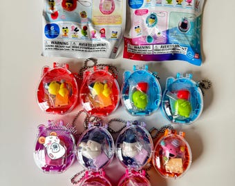 May include: A collection of Tamagotchi Collectibles, featuring various colorful, pocket-sized virtual pet toys. Each toy is encased in a clear, oval-shaped container with a keychain. The packaging includes the text "Tamagotchi Collectibles".