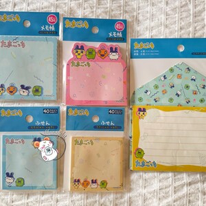 May include: Assortment of Tamagotchi-themed stationery, including memo pads and a letter set. The memo pads come in various colors like blue, pink, and beige, with cute character illustrations. The letter set includes an envelope and lined paper.