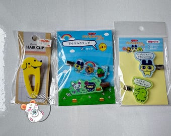 May include: Three packaged hair clips. One is a yellow dog-shaped hair clip with the text "HAIR CLIP". The other two are acrylic clips with cartoon characters and Japanese text. One clip has a rainbow design.