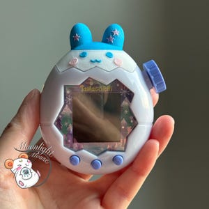 May include: A white Tamagotchi digital pet device featuring a blue rabbit-ear top attachment with pink star accents. The screen frame has a winter-themed pattern with trees and snowflakes. It includes three round blue buttons and a side dial.