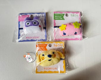 May include: Three mascot hair ties in clear packaging. The top left features a white and purple character with the text "MASKUTCHI". The top right shows a pink character with yellow flowers and the text "FunnWHICHI". The bottom shows a yellow dog with the text "POCHITCHI".