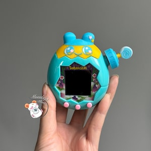 May include: A teal Tamagotchi virtual pet toy with blue accents, yellow highlights, and pink buttons. The screen displays a black rectangle. The toy has a lollipop-shaped antenna and the text "Tamagotchi".