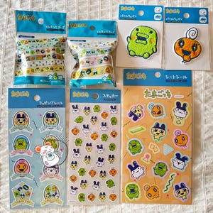 May include: A selection of Japanese character merchandise, including sticker sheets, patches, and masking tape rolls. The items feature cartoon characters in bright colours, with Japanese text on the packaging. The items are arranged on a white surface.