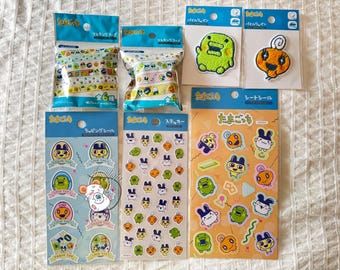 May include: A collection of colorful Japanese character merchandise, including sticker sheets, patches, and masking tape rolls. The items feature various cartoon characters in bright colors, with Japanese text on the packaging. The items are arranged on a white surface.