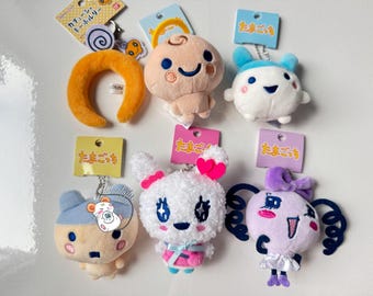 May include: A collection of plush keychains featuring various cartoon characters. The keychains are in different colors, including orange, blue, white, and pink. Each keychain has a tag with Japanese text.