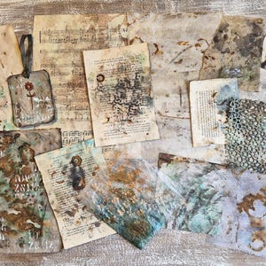 May include: A collection of assorted ephemera including aged music sheets, stained paper scraps, a small rectangular tag with a flower illustration, and a piece of green netting. The items feature distressed textures, brown ink stains, and numbers.