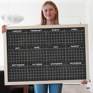 May include: A black chalkboard calendar with a wooden frame. The calendar is divided into twelve months, with each month having its own grid of squares. The calendar is being held by a person.