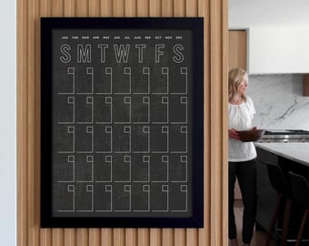 May include: A black framed calendar with a white grid and the days of the week labeled "SMTWTFS". The calendar is hanging on a wall with wood paneling.