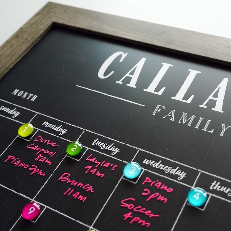 May include: Close-up of a black framed family calendar chalkboard with the words 'Calla Family' at the top.  The chalkboard features a monthly grid with days of the week labeled. Colorful magnetic markers are placed on various days, indicating scheduled events written in pink.  Events include 'Drive Carpool 8am,' 'Layla's 9am,' 'Brunch 11am,' 'Piano 2pm,' and 'Soccer 4pm.'  The numbers 1 through 4 and 9 are visible on the magnetic markers.