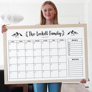 May include: A white dry-erase calendar with a wooden frame. The calendar has a monthly grid with days of the week labeled. The top of the calendar says "The Lockett Family" and has a mountain design on each side. The calendar also has a section for a menu and notes.