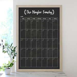 May include: A wooden framed chalkboard with a black surface and white grid lines. The chalkboard has the text "The Naylor Family" with a decorative leaf design on either side of the text. The chalkboard is a monthly calendar with the days of the week labeled.
