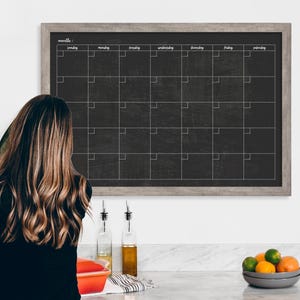 May include: A black chalkboard calendar with a gray wood frame hangs on a white wall. The calendar has the days of the week listed across the top and the numbers of the month listed down the side. The calendar is blank, ready for someone to write in their appointments.