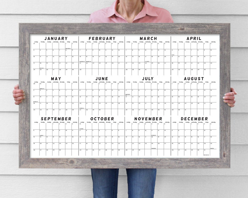 May include: A large, rectangular calendar with a gray wooden frame. The calendar displays all twelve months, with each month's name in bold black text. The calendar grid is black on a white background.