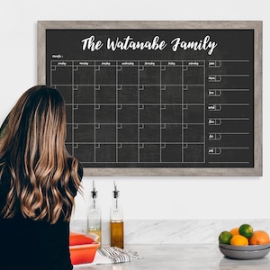 May include: A black chalkboard with a white frame and the text "The Watanabe Family" at the top. The chalkboard is divided into a calendar grid with days of the week labeled and a section for notes.
